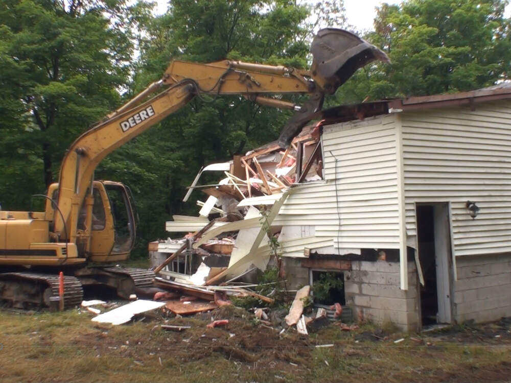 A demolition contractor uses a hydraulic excavator to take a bite out of a wood frame house structure during whole-home demolition and disposal in Muskoka, Ontario.