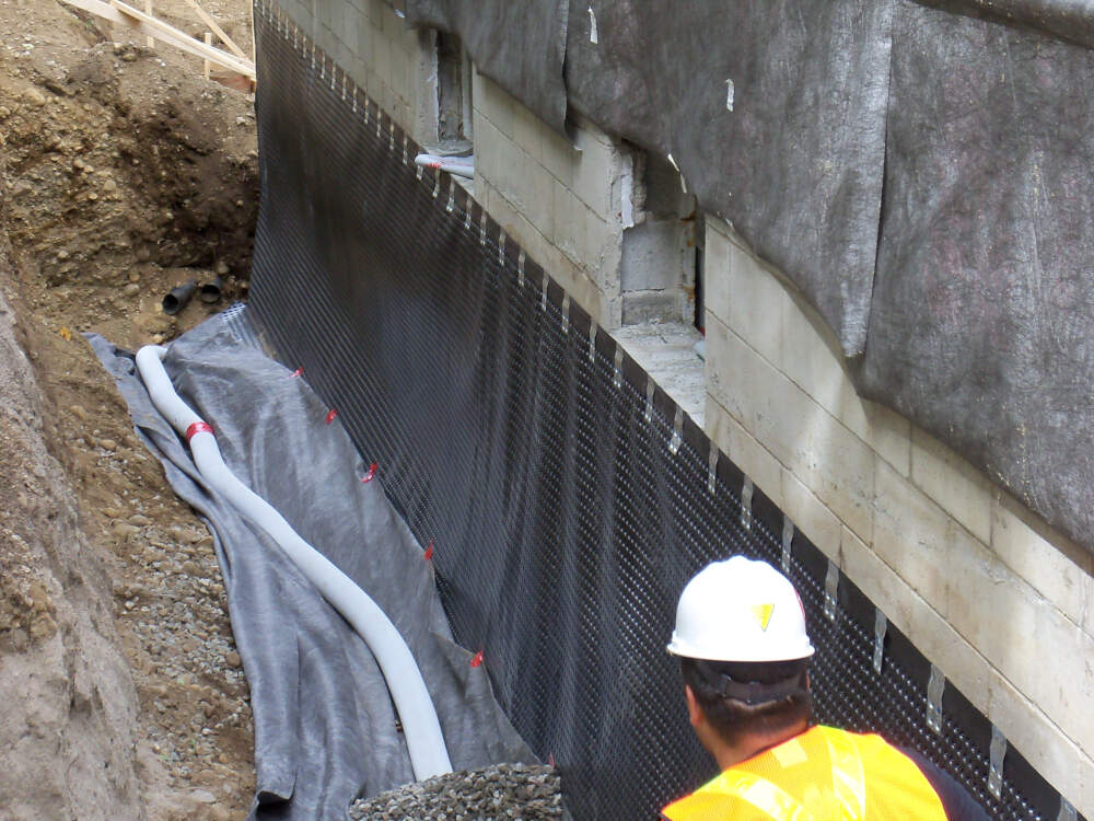 Foundation drainage sheet, drainage piping and gravel drainage aggregate are installed by an apprentice carpenter wearing a reflective safety vest in this Muskoka construction site image.