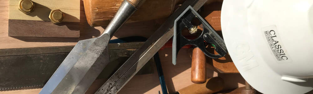 Paring chisel, combination square, a hand block plane, a dovetail saw and other carpentry tools are pictured beside a Classic Muskoka Homes branded hard hat.